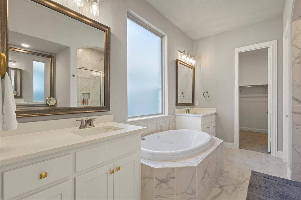 Dual vanities with white cabinetry, quartz-style countertops, drop-in soaking tub, tile surround, and large framed mirrors
