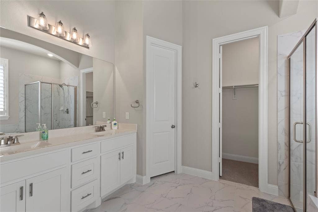 Double vanity featuring white cabinetry, a neutral solid-surface countertop, full-width mirror, five-light fixture, and walk-in shower with stone-patterned tile surround