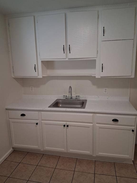 White cabinetry featuring matte black hardware, integrated storage shelving, solid-surface countertops, stainless steel drop-in sink with chrome faucet, and tiled flooring