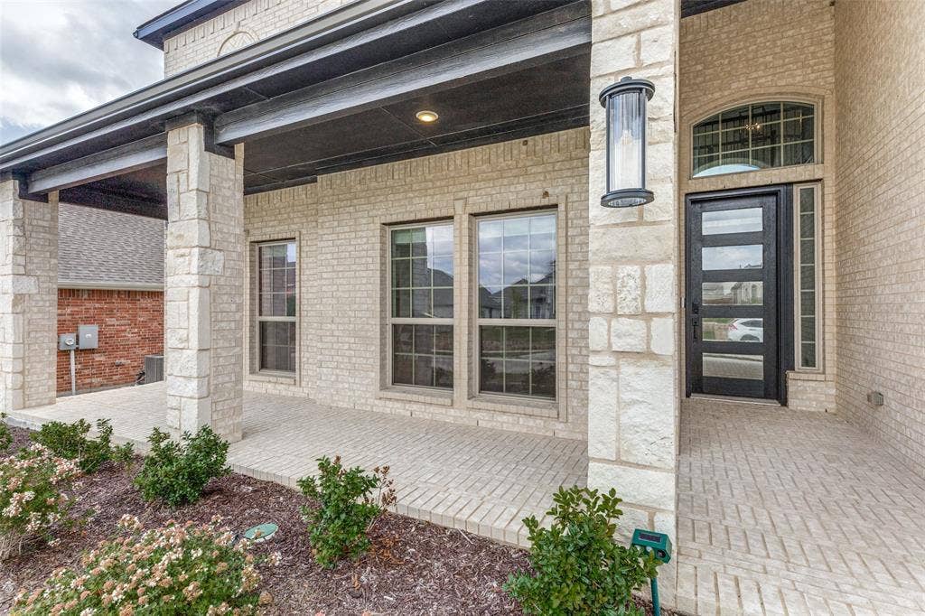 Covered front porch featuring light-toned brick masonry, stacked stone support columns, a dark-framed multi-lite front door with arched transom, and integrated wall-mounted exterior lighting