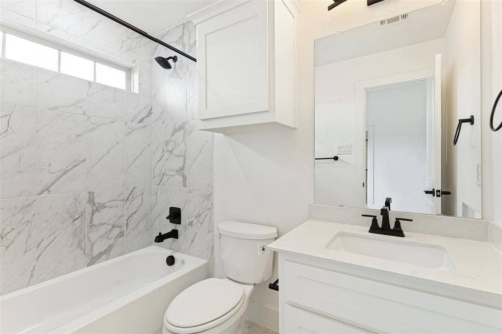 Bathroom featuring a shower-tub combination with stone-patterned wall tile, a white vanity with a light-toned countertop, a rectangular undermount sink, matte black hardware, and an overhead cabinet