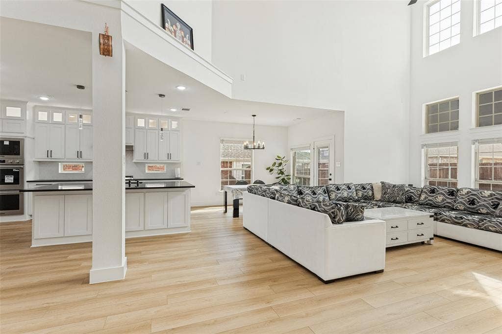 Open-concept interior featuring double-height ceilings, wood-finish flooring, white shaker-style cabinetry, a built-in double oven, and multiple tiered windows