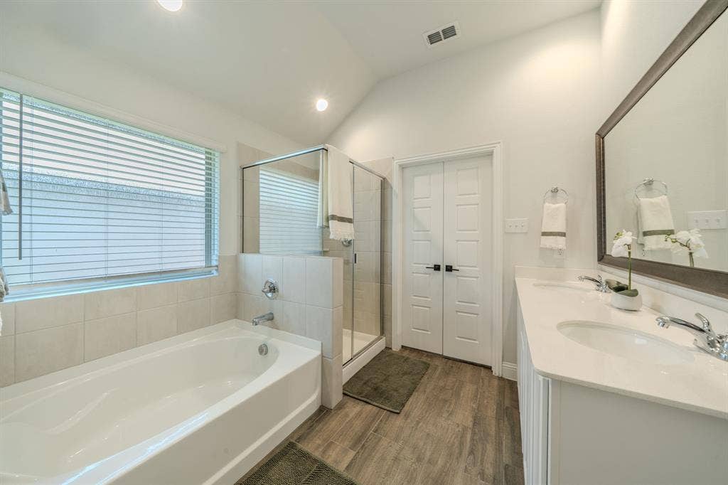 Bathroom featuring a vaulted ceiling, dual-sink vanity with light-toned countertop, glass-enclosed shower, built-in soaking tub, and wood-finish flooring
