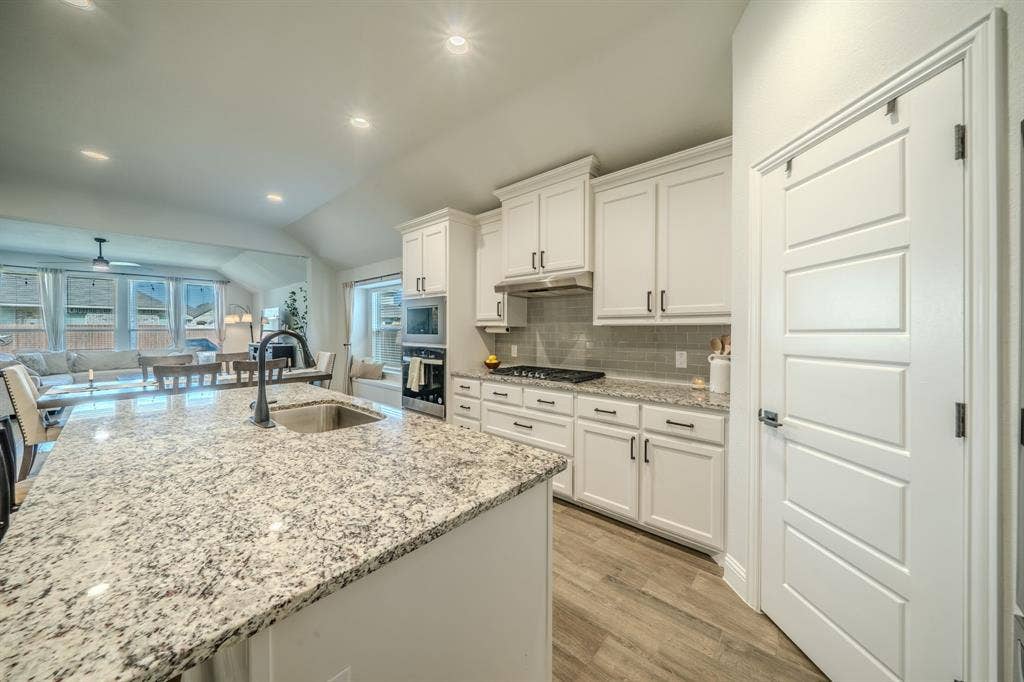 Kitchen featuring a central island with a stone-finish countertop and undermount sink, white cabinetry, a built-in oven, a gas cooktop, and wood-finish flooring