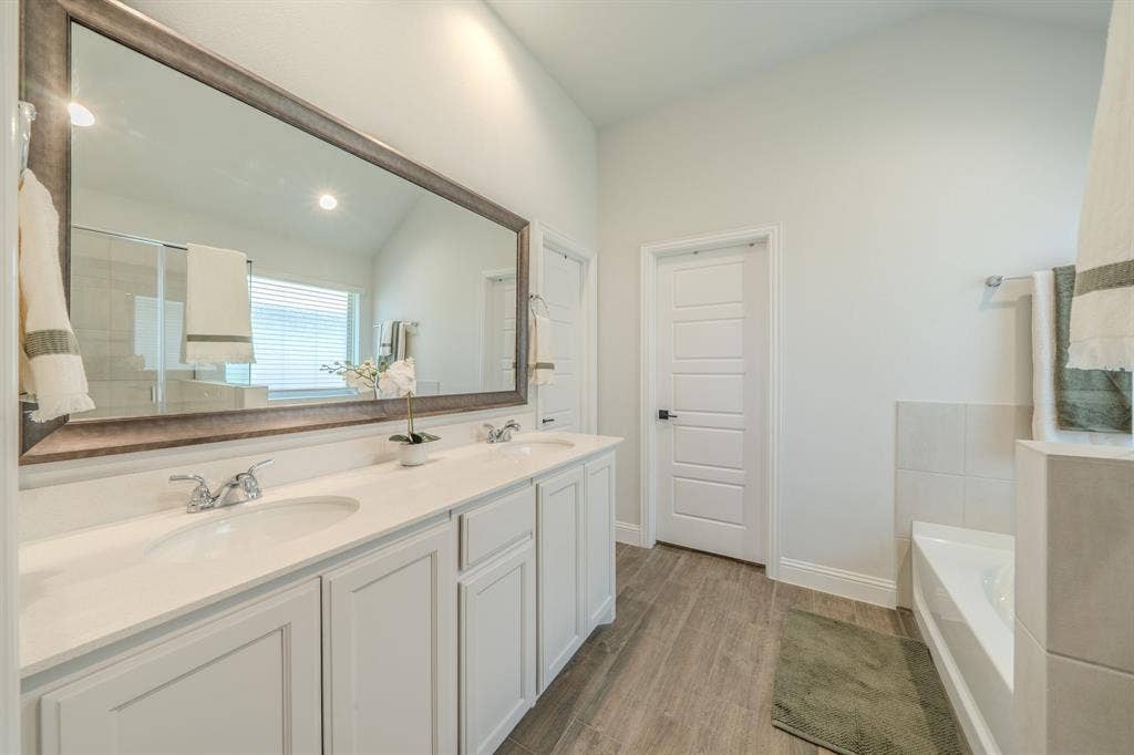 Dual-sink vanity with white cabinetry and solid surface countertop, large framed mirror, soaking tub with tiled surround, and wood-finish flooring