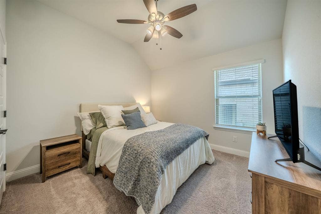 Vaulted ceiling with a central multi-blade ceiling fan and light fixture, neutral carpeted flooring, and a single window with integrated blinds