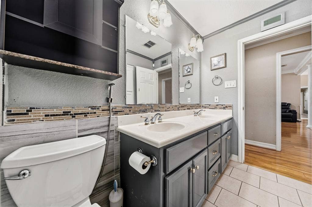 Dual-sink vanity with dark cabinetry, stone-finish backsplash, neutral tile flooring, and integrated wall mirrors