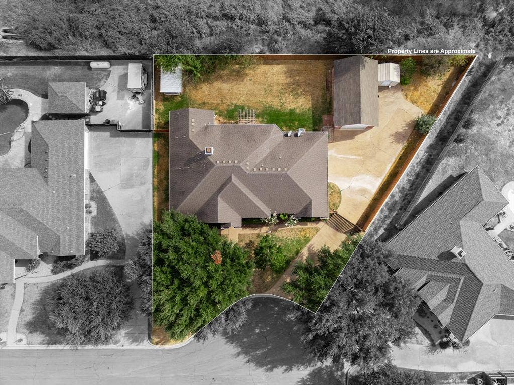 Aerial perspective of a property featuring a multi-pitched shingle roof, expansive paved driveway, detached garage structure, fenced rear lot, and mature trees