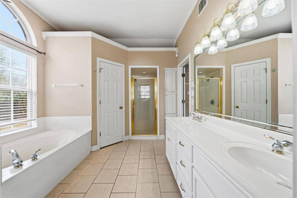 Bathroom featuring tile flooring, dual-basin white vanity with expansive mirror, recessed garden tub, and a separate glass-enclosed shower