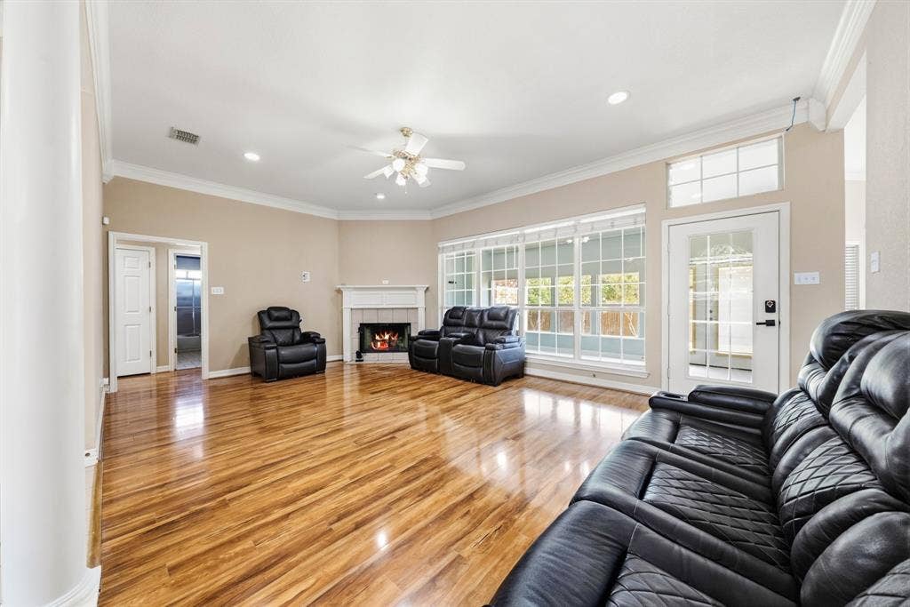 Living space featuring wood-finish flooring, a traditional fireplace with a white mantel, crown molding, a ceiling fan, and large windows with transom detailing