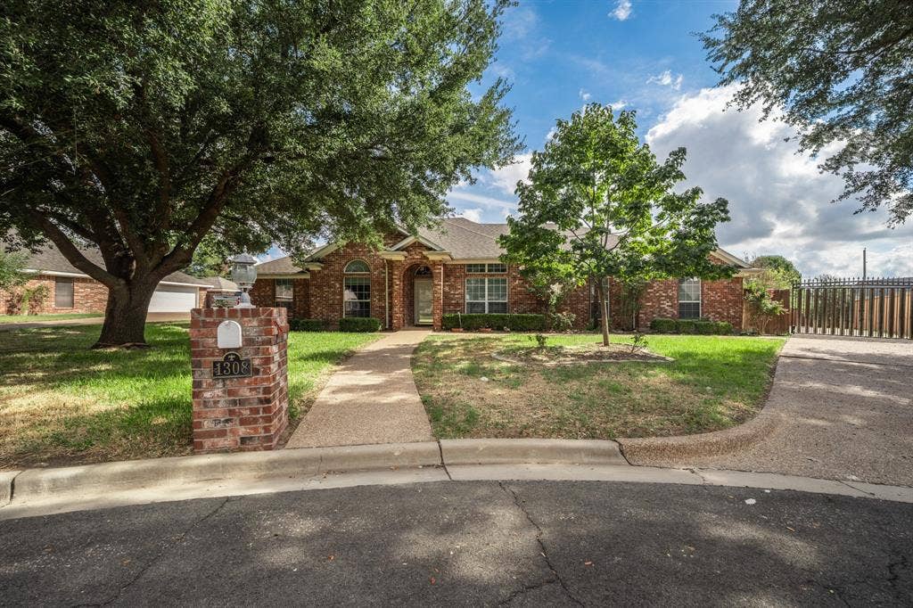 Single-story brick exterior featuring a recessed arched entryway, a stone-aggregate walkway, a matching brick mailbox, and mature shade trees