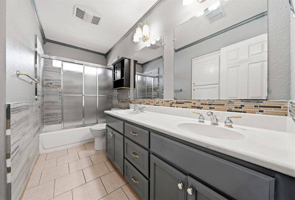 Dual-sink vanity with gray cabinetry and solid-surface countertop, full-width wall mirror, mosaic tile backsplash, and a tiled shower-tub combination with sliding glass doors