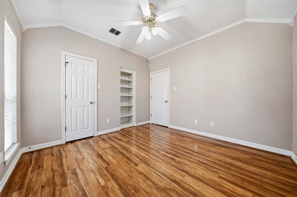 Vaulted ceiling featuring a central multi-blade ceiling fan, wood-finish flooring, built-in recessed shelving, and two six-panel interior doors