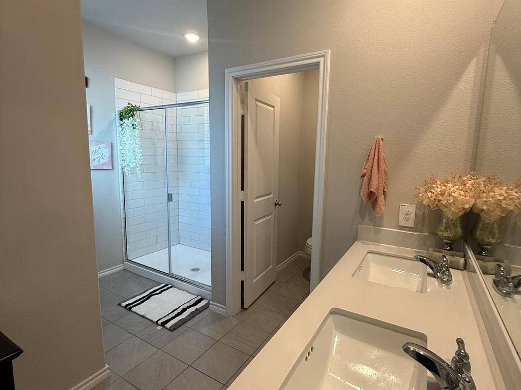 Bathroom featuring a dual-sink vanity with solid-surface countertop, walk-in shower with white subway tile surround, and neutral floor tile