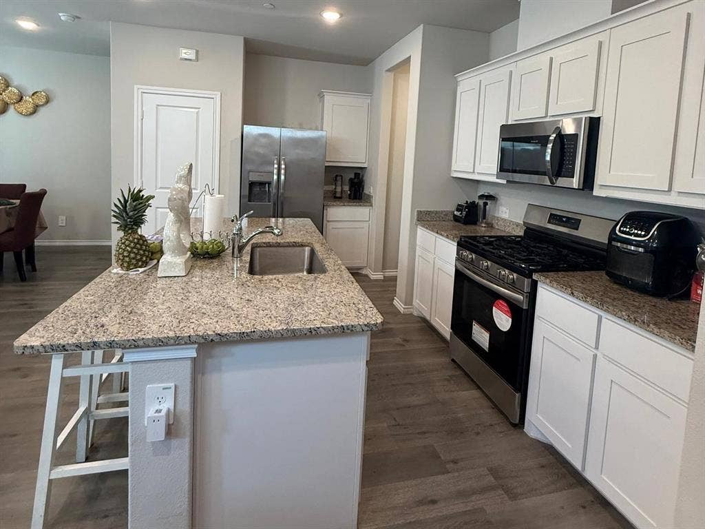 Kitchen featuring white shaker-style cabinetry, stone-finish countertops, stainless steel appliances, a center island with an integrated sink, and wood-finish flooring