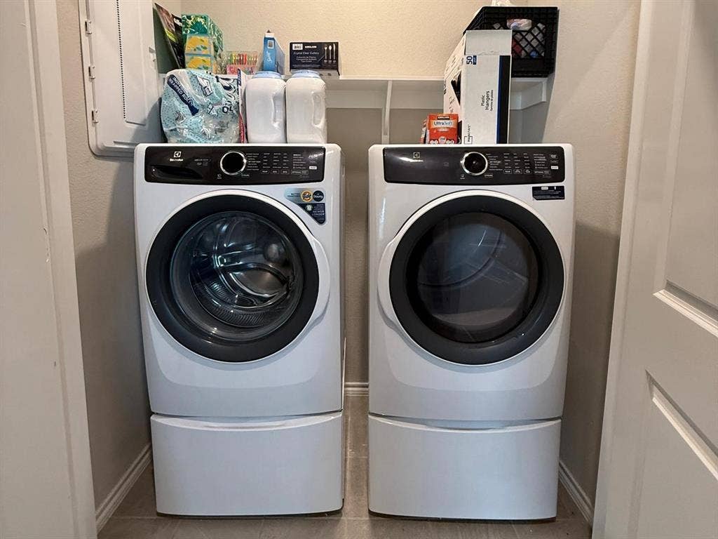 Dedicated laundry space featuring high-capacity front-load washer and dryer set, built-in overhead storage shelving, and electrical panel access