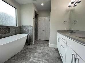Freestanding soaking tub with floor-mounted faucet, vanity with dual undermount sinks, stone-finish tile flooring, recessed wall niche, and multi-light vanity fixture