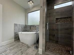 Freestanding soaking tub with a floor-mounted faucet, walk-in shower with stone tile flooring and recessed shelving, wood-finish tile flooring, and a crystal chandelier