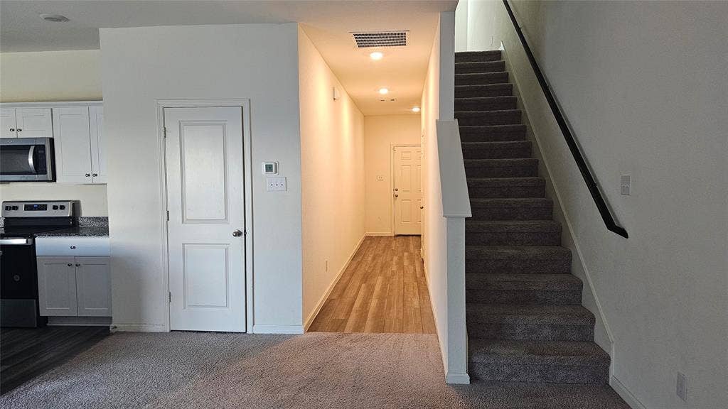Kitchen area featuring white cabinetry, stone-finish countertops, and stainless steel appliances, complemented by wood-finish flooring and carpeted staircase with a dark handrail