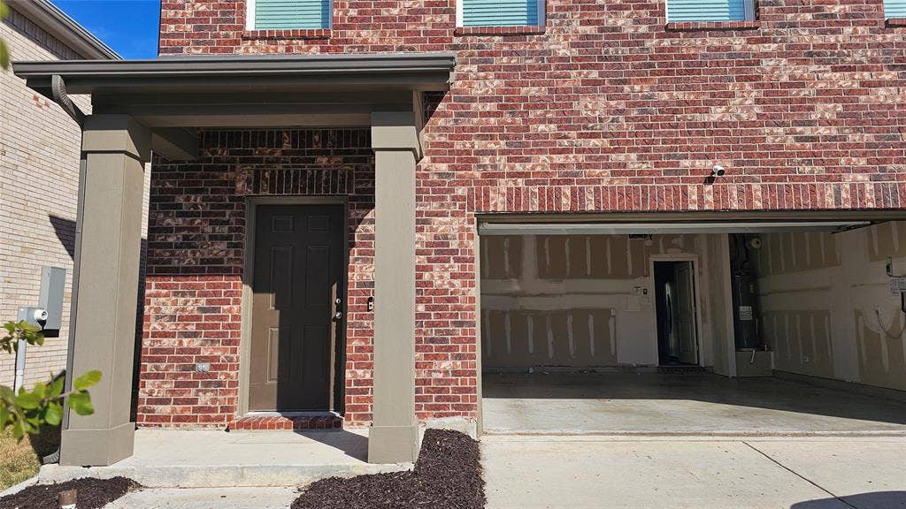 Exterior featuring multi-tonal brick masonry, a covered porch with concrete foundation, two structural pillars, and a concrete driveway leading to a two-car garage