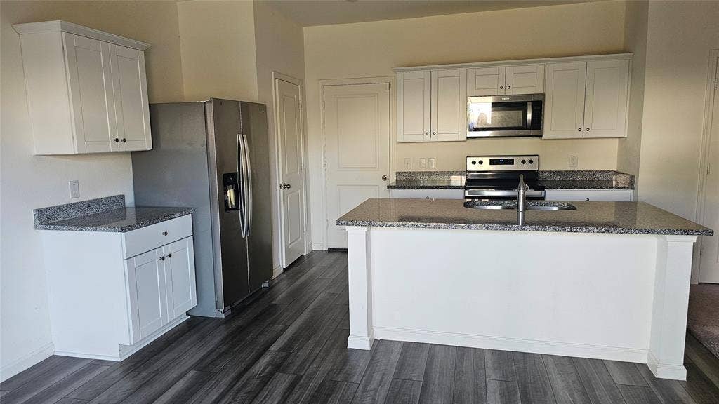 Kitchen featuring white shaker-style cabinetry, speckled stone-finish countertops, stainless steel appliances, a center island with a dual-basin sink, and wood-finish flooring