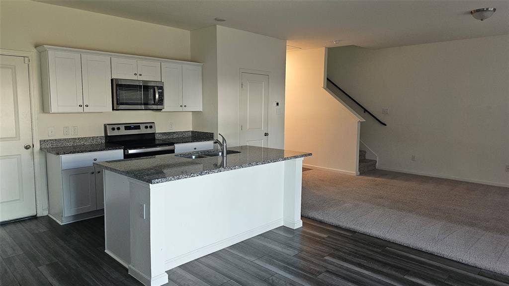 Kitchen featuring white cabinetry, stone-finish countertops, stainless steel appliances, a center island with a dual-basin sink, and wood-finish flooring