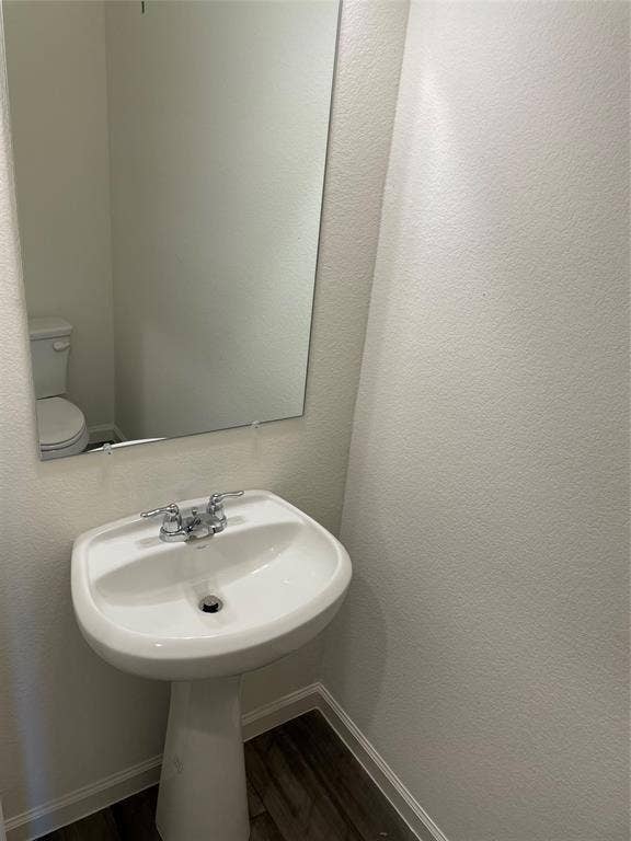 White pedestal sink with a dual-handle chrome faucet, large rectangular wall-mounted mirror, white ceramic toilet, and wood-finish flooring