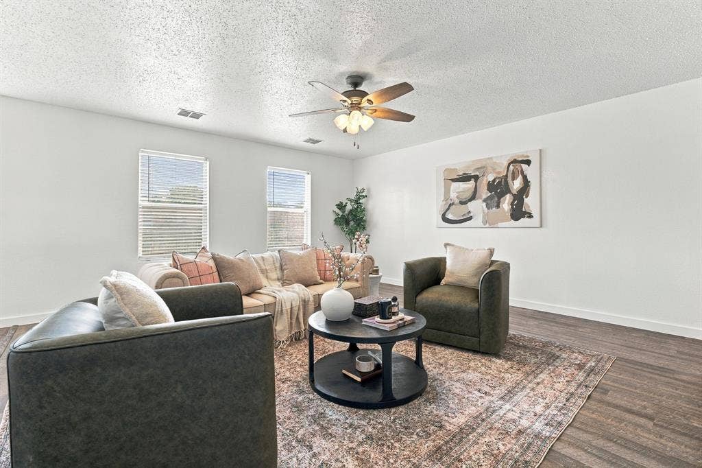 Living area featuring wood-finish flooring, two dual-pane windows with horizontal blinds, and a central ceiling fan with integrated lighting
