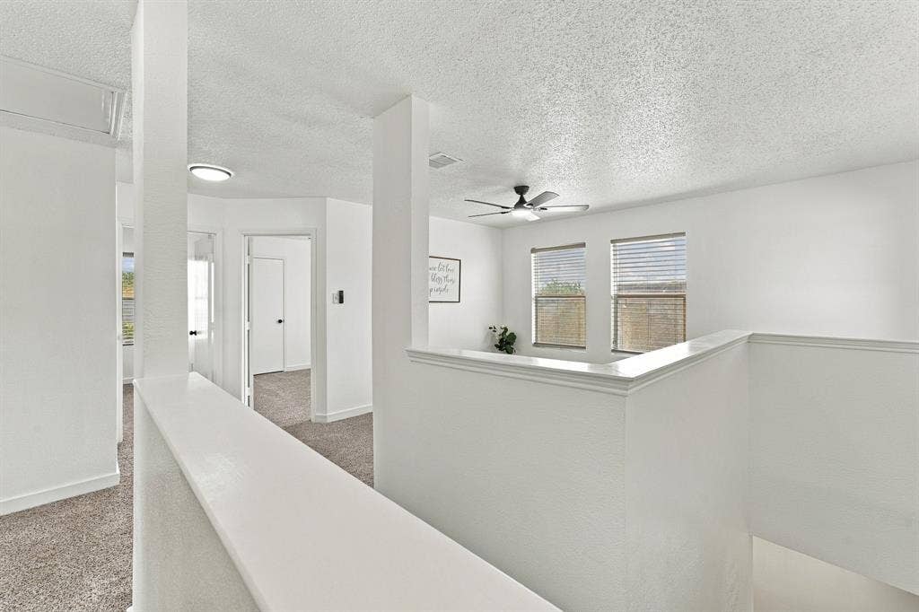 Open floor plan interior featuring textured ceilings, carpeted flooring, a multi-blade ceiling fan with integrated lighting, and dual windows with horizontal blinds