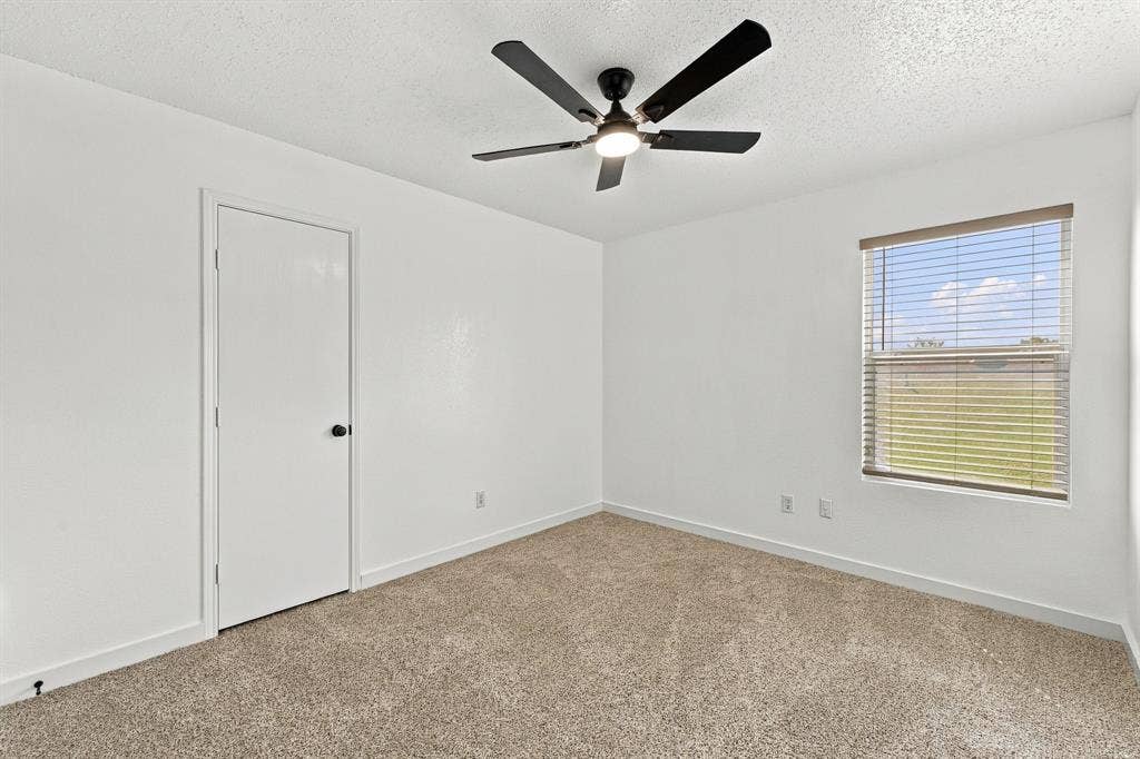 Neutral-toned carpeting, matte black ceiling fan with integrated lighting, single-panel interior door, and window with horizontal blinds