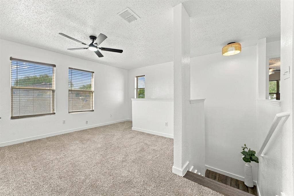 Neutral-toned wall-to-wall carpeting, three multi-pane windows with horizontal blinds, a matte black multi-blade ceiling fan, and a woven drum light fixture