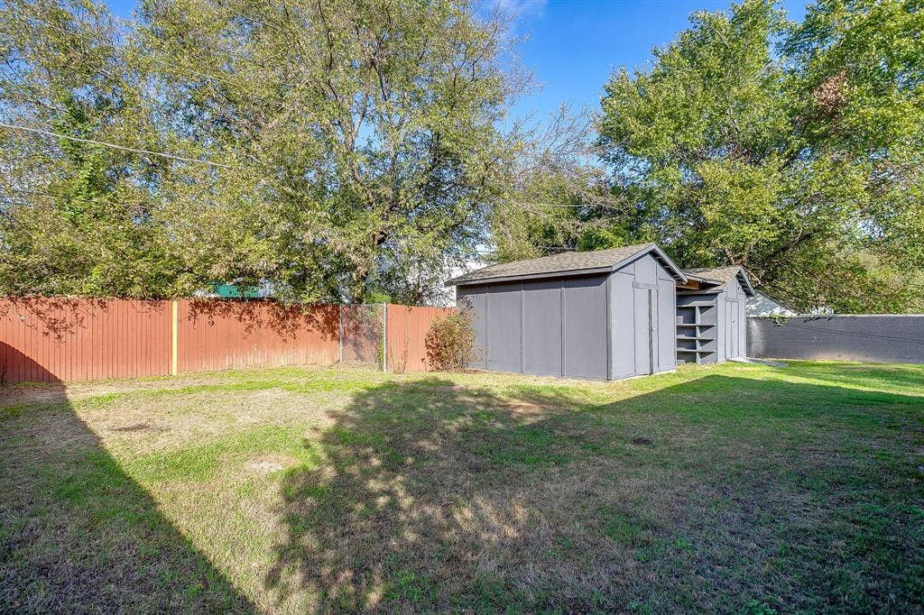 Backyard featuring a grass lawn, mature trees, a corrugated metal perimeter fence, and a detached storage structure with integrated exterior shelving