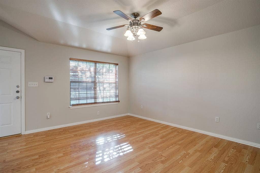 Interior room featuring wood-finish flooring, vaulted ceiling, multi-blade ceiling fan with integrated lighting, and window with horizontal blinds