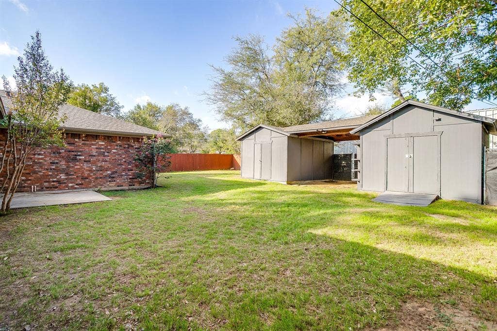 Expansive backyard featuring a brick exterior facade, a concrete patio, two storage structures, and perimeter wood fencing