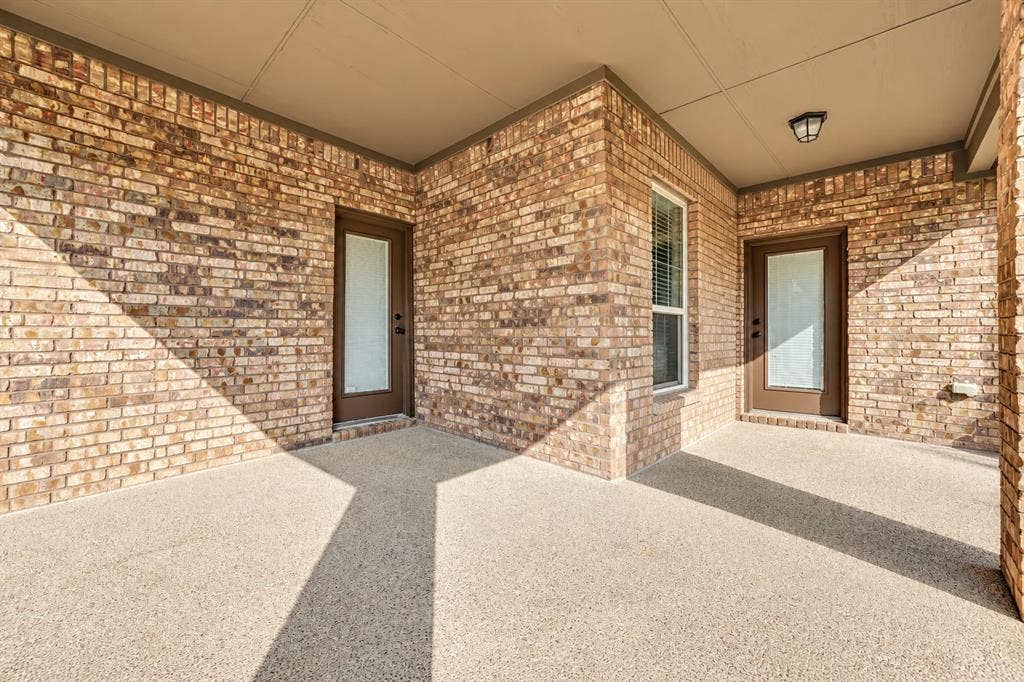 Covered patio featuring red brick masonry, aggregate concrete flooring, two glass-paneled exterior doors, and a flush-mount overhead light fixture