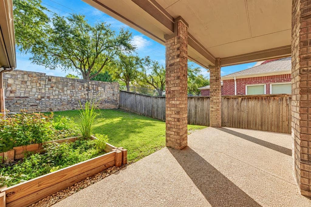 Covered patio featuring concrete flooring and brick columns, complemented by a stone perimeter wall, wooden raised garden bed, and grass lawn