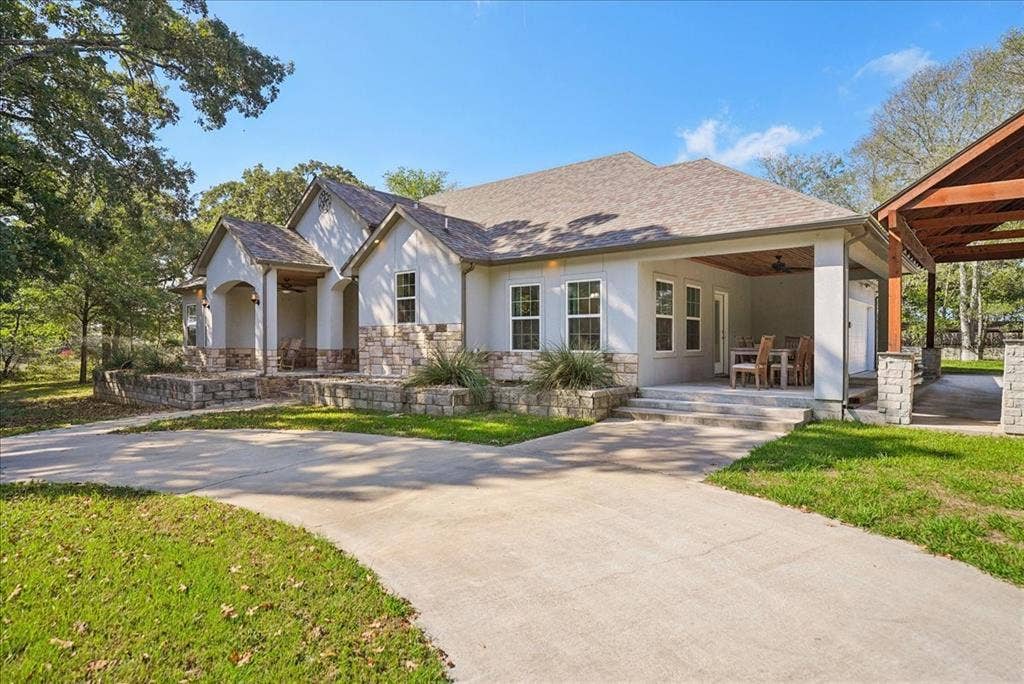 7320 County Road 3512, Quinlan, TX 75474 Single-story exterior featuring a light-colored masonry facade with stone veneer accents, a concrete driveway, a covered patio area with a wood-plank ceiling, and a detached wooden pergola structure