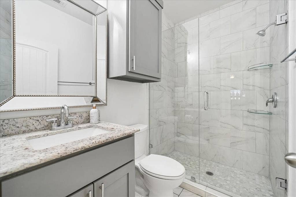 7320 County Road 3512, Quinlan, TX 75474 Bathroom featuring a granite vanity countertop, gray shaker-style cabinetry, a framed wall mirror, a glass-enclosed shower with stone-tile surround, and a pebble-stone shower floor