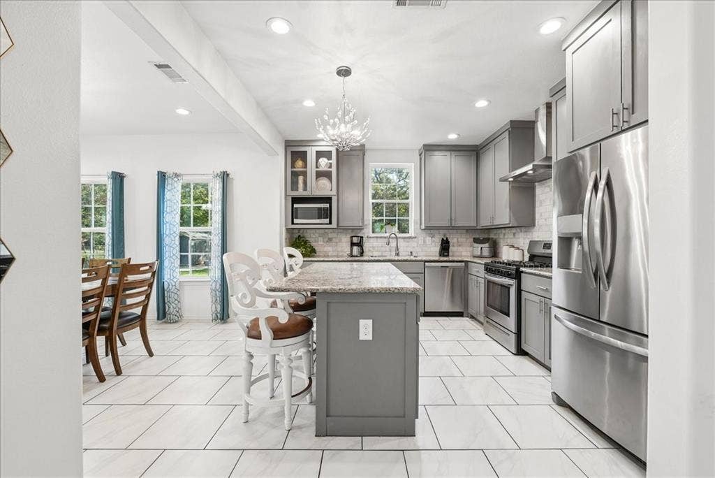 7320 County Road 3512, Quinlan, TX 75474 Kitchen featuring gray shaker-style cabinetry, a central island with stone-surface countertop, stainless steel appliances, a light-toned tile backsplash, and decorative chandelier lighting
