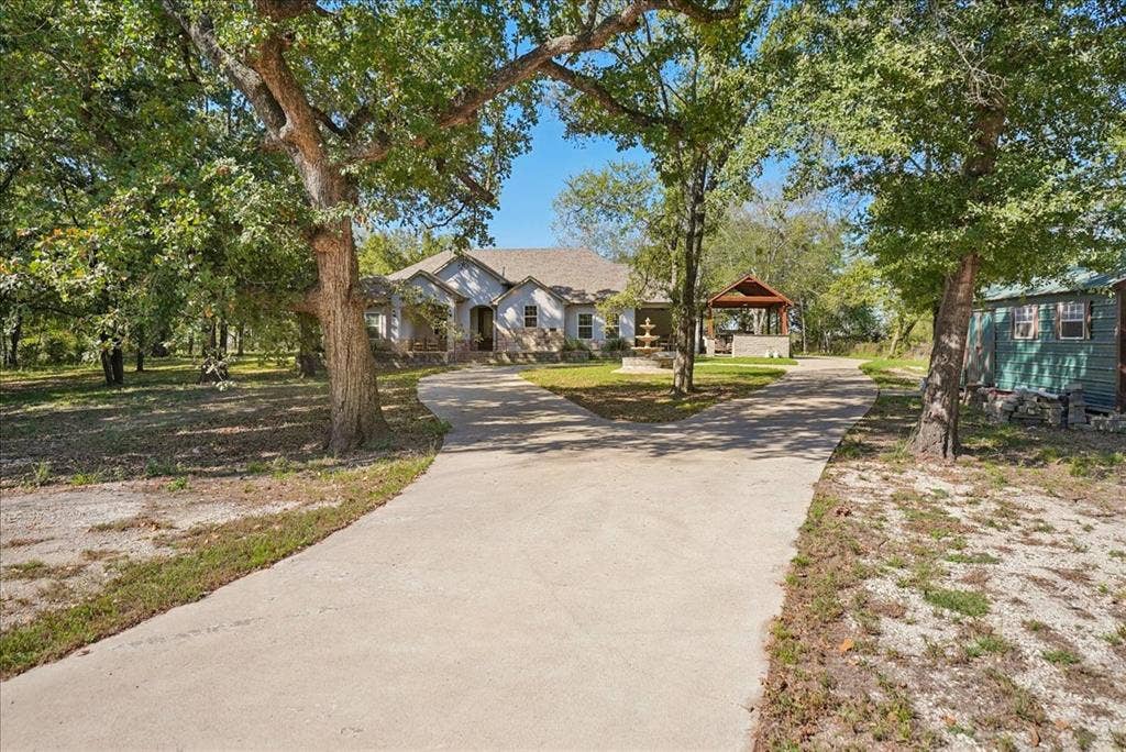 7320 County Road 3512, Quinlan, TX 75474 Single-story residence featuring a bifurcated concrete driveway, stone and neutral-tone siding exterior, decorative water feature, and a detached wooden-frame outdoor pavilion situated on a lot with mature trees