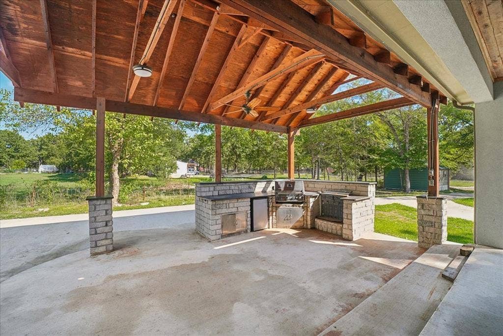 7320 County Road 3512, Quinlan, TX 75474 Covered outdoor kitchen featuring a timber-framed vaulted ceiling, stone pillar supports, stainless steel built-in grill, integrated refrigeration, and masonry countertop
