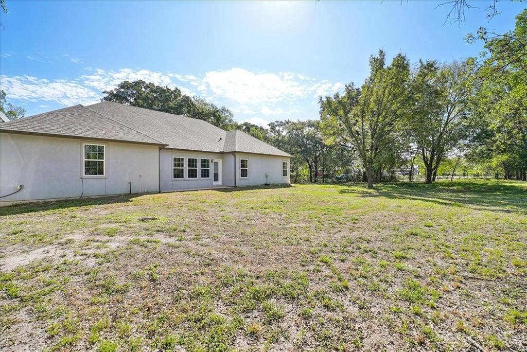 7320 County Road 3512, Quinlan, TX 75474 Exterior rear elevation featuring a light-toned stucco facade, a multi-hip roofline, and expansive grounds with mature trees