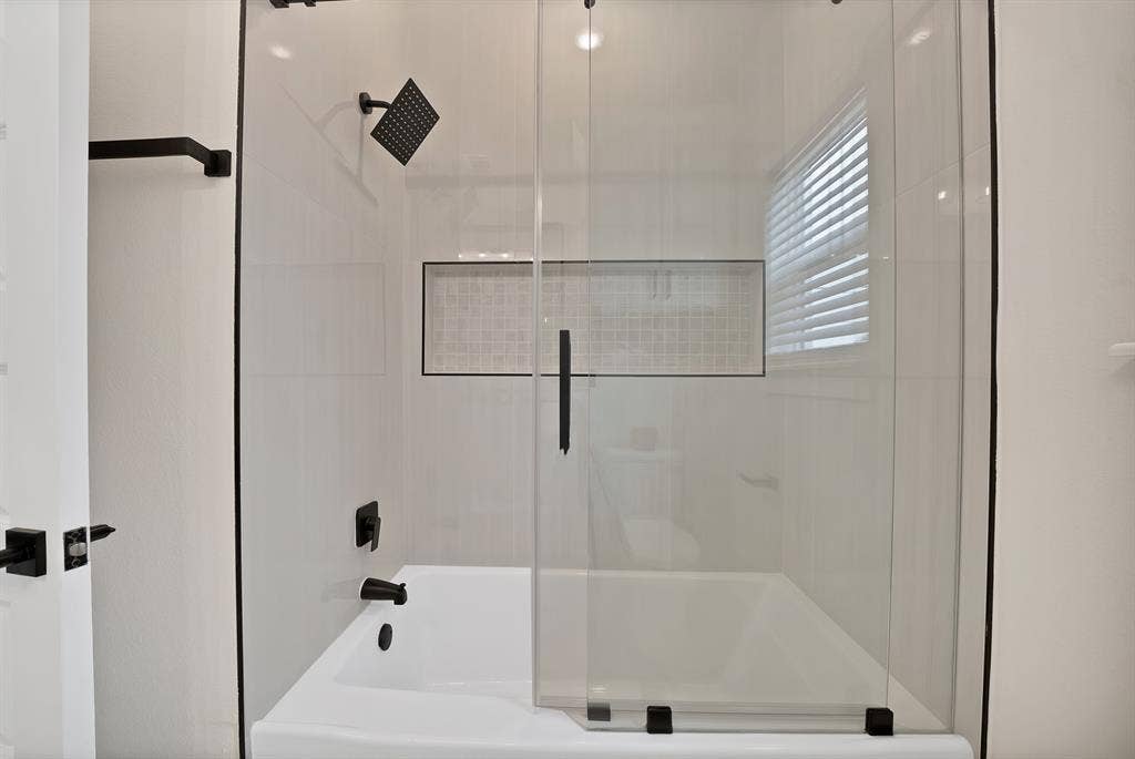 Bathtub and shower combination featuring white tiled surround, built-in recessed tiled niche, sliding glass door, and matte black hardware