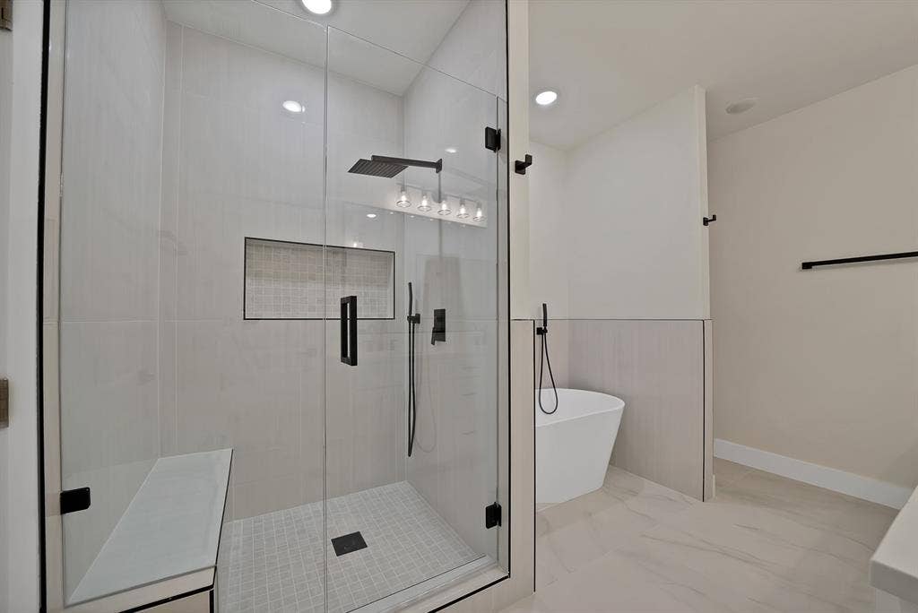 Walk-in shower featuring a frameless glass enclosure, built-in tiled bench, recessed niche, and matte black rainfall shower head, complemented by a freestanding soaking tub