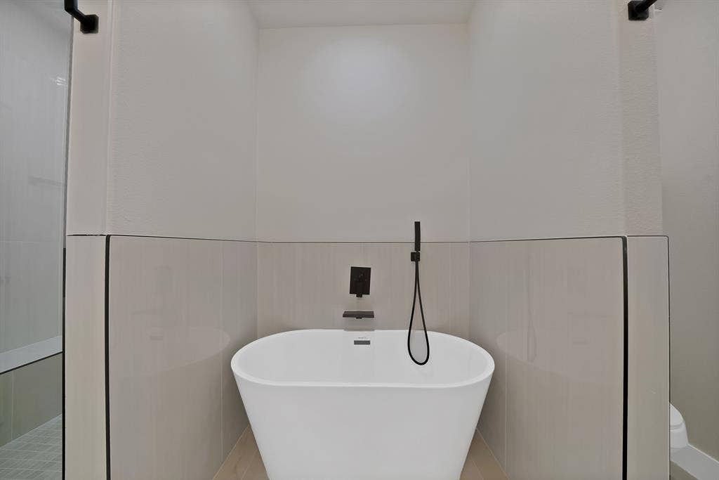 Freestanding white soaking tub with matte black wall-mounted faucet and handheld shower sprayer, complemented by floor-to-ceiling neutral tile surround