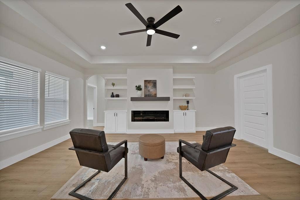 Living area featuring a tray ceiling, wood-finish flooring, built-in shelving, white cabinetry, and a linear fireplace with a mantel