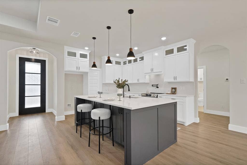 Kitchen featuring a central island with a contrasting dark base, white cabinetry with glass-front uppers, light-toned wood-finish flooring, quartz countertops, and three minimalist pendant lights