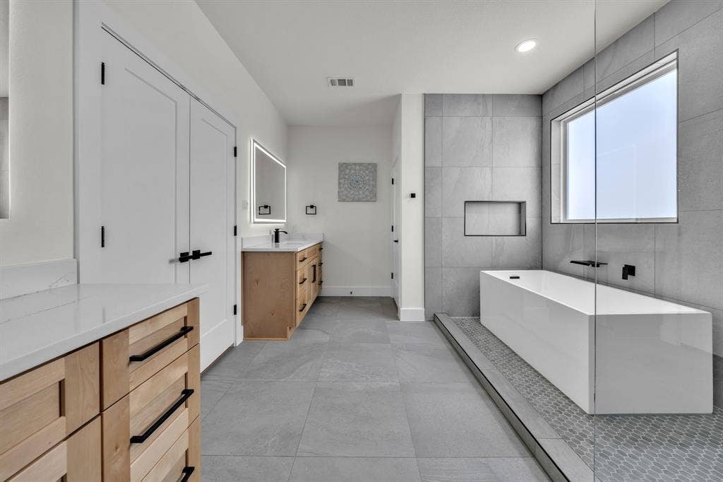 3639 Pueblo Street, Dallas, TX 75212 Bathroom featuring a freestanding rectangular soaking tub, large-format grey floor and wall tile, dual wood-finish cabinetry with white stone countertops, and matte black hardware