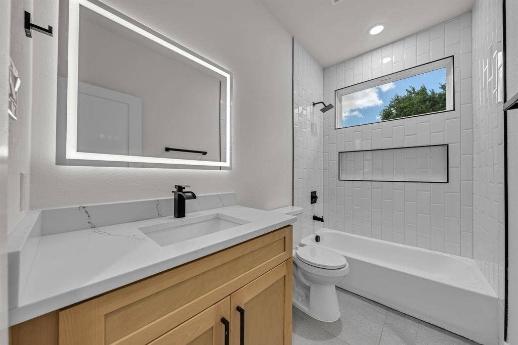 3639 Pueblo Street, Dallas, TX 75212 Bathroom featuring a vanity with wood-finish cabinetry, white countertop, integrated rectangular sink, and matte black faucet, complemented by an LED-backlit mirror, subway-tiled tub surround with a recessed niche, and horizontal transom window