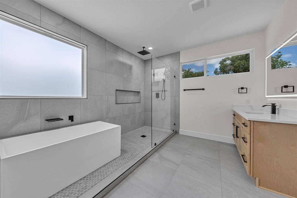 3639 Pueblo Street, Dallas, TX 75212 Bathroom featuring a freestanding soaking tub, walk-in glass-enclosed shower with a built-in niche, light wood-finish vanity with a stone countertop, large-format floor tiling, and matte black fixtures